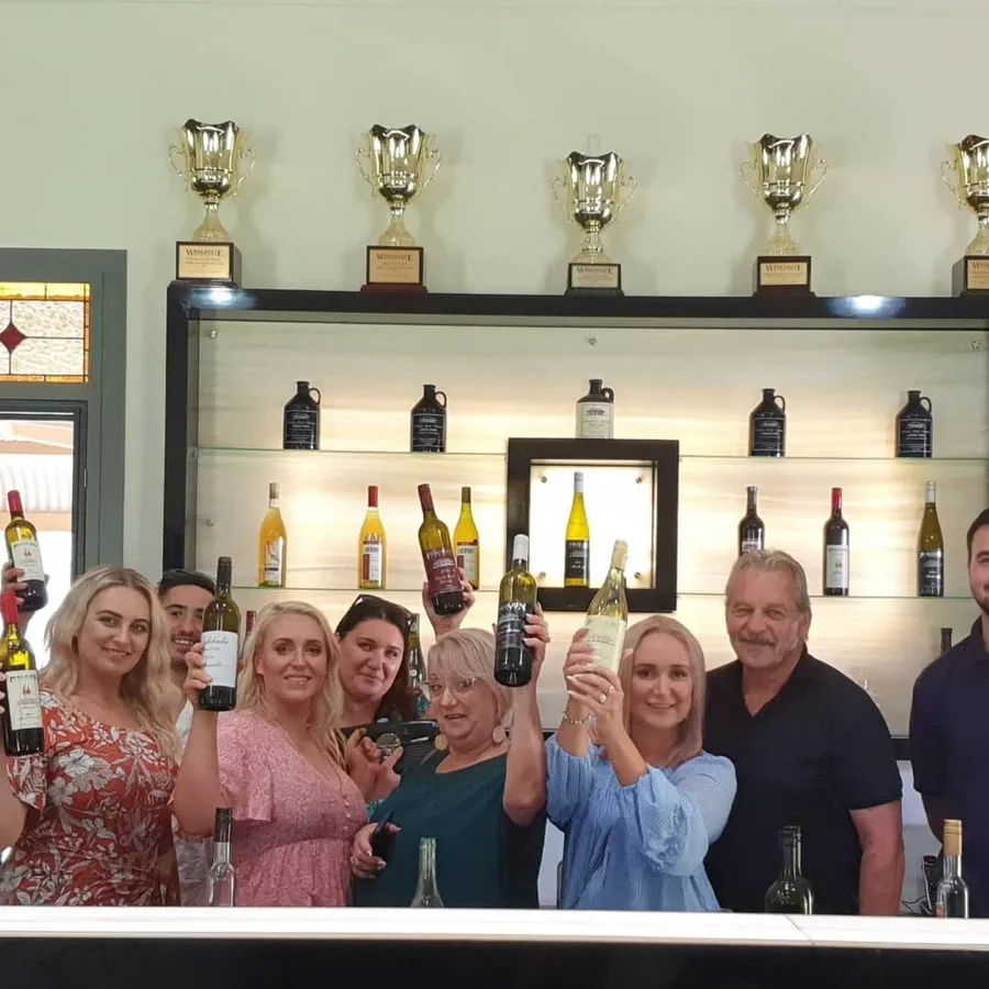 Guests hold up their bottles in front of a shelf of wine and trophies