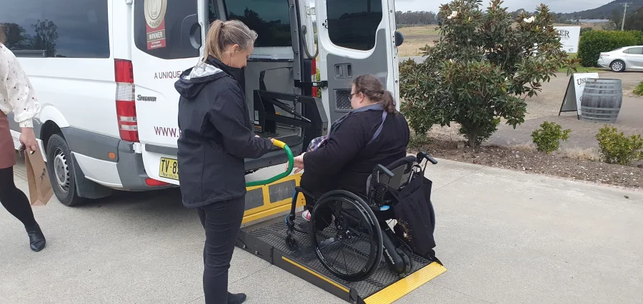 A team member guides a guest in a wheelchair onto the minibus hydraulic lift, vineyard behind them