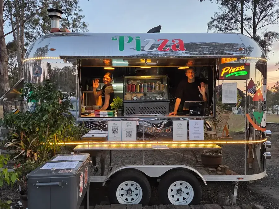 The Just One More Pizza van parked among gum trees, its service window open with two staff inside